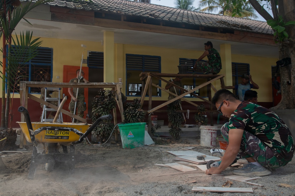 A construction or renovation project is underway outside a yellow building, likely in Southeast Asia. Multiple workers in military camouflage uniforms and civilian clothing are engaged in various construction tasks. The scene includes scaffolding, tools, and building materials scattered across a sandy work area.