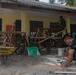 A construction or renovation project is underway outside a yellow building, likely in Southeast Asia. Multiple workers in military camouflage uniforms and civilian clothing are engaged in various construction tasks. The scene includes scaffolding, tools, and building materials scattered across a sandy work area.