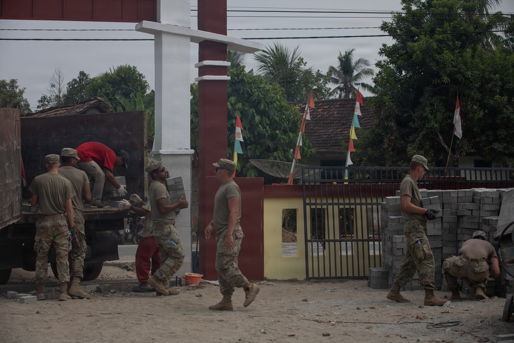 Military personnel in camouflage uniforms are engaged in construction or civic action work at what appears to be a compound entrance in a tropical Southeast Asian location. Soldiers are unloading paving stones or concrete blocks from a truck and laying them on the ground. The presence of Indonesian flags and tropical vegetation suggests this is likely a humanitarian assistance or civil-military cooperation project in Indonesia.