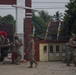 Military personnel in camouflage uniforms are engaged in construction or civic action work at what appears to be a compound entrance in a tropical Southeast Asian location. Soldiers are unloading paving stones or concrete blocks from a truck and laying them on the ground. The presence of Indonesian flags and tropical vegetation suggests this is likely a humanitarian assistance or civil-military cooperation project in Indonesia.