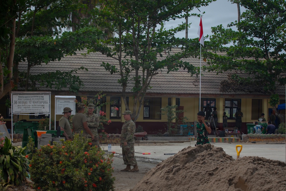 The image shows military personnel, appearing to be a mix of U.S. soldiers in MultiCam/OCP uniforms and an Indonesian soldier in green camouflage, gathered at what is identified as SD Negeri 12 Martapura school in Kecamatan Martapura, Indonesia. A pile of sand and construction materials suggest an ongoing civil engineering or humanitarian assistance project at the school compound. The Indonesian national flag (Merah Putih - red and white) is visible on a flagpole in the background.
