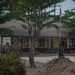 The image shows military personnel, appearing to be a mix of U.S. soldiers in MultiCam/OCP uniforms and an Indonesian soldier in green camouflage, gathered at what is identified as SD Negeri 12 Martapura school in Kecamatan Martapura, Indonesia. A pile of sand and construction materials suggest an ongoing civil engineering or humanitarian assistance project at the school compound. The Indonesian national flag (Merah Putih - red and white) is visible on a flagpole in the background.