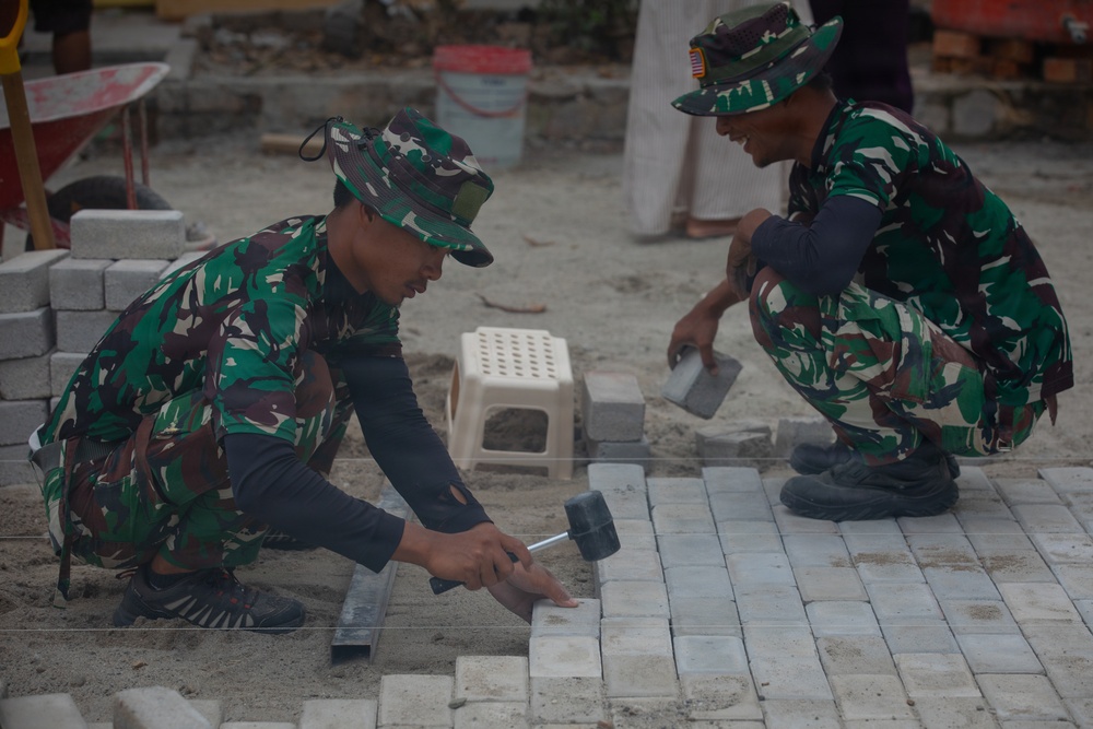 Two military personnel in camouflage uniforms are laying interlocking paving stones or cobblestones on a sandy surface. One soldier is using a rubber mallet and a metal straightedge to set and align the pavers, while the other assists by holding what appears to be a small container of sand or mortar. The setting appears to be a construction or reconstruction site with building materials visible in the background.