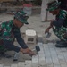 Two military personnel in camouflage uniforms are laying interlocking paving stones or cobblestones on a sandy surface. One soldier is using a rubber mallet and a metal straightedge to set and align the pavers, while the other assists by holding what appears to be a small container of sand or mortar. The setting appears to be a construction or reconstruction site with building materials visible in the background.