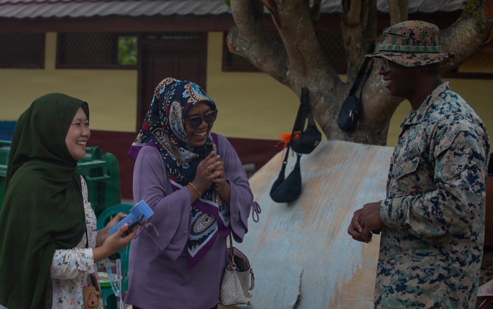 A U.S. Marine in digital camouflage uniform and cover is engaged in a friendly conversation with two women wearing hijabs in what appears to be an outdoor community or school setting. The interaction appears warm and jovial, with the women visibly laughing. The setting suggests a civil engagement or humanitarian assistance event.