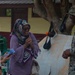 A U.S. Marine in digital camouflage uniform and cover is engaged in a friendly conversation with two women wearing hijabs in what appears to be an outdoor community or school setting. The interaction appears warm and jovial, with the women visibly laughing. The setting suggests a civil engagement or humanitarian assistance event.