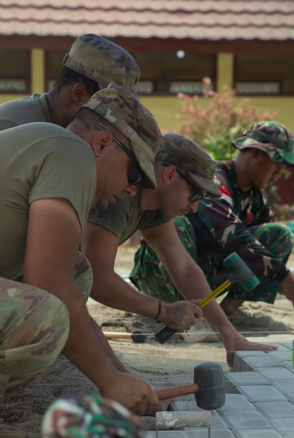 A group of military personnel in mixed camouflage uniforms are collaboratively laying interlocking paving stones or bricks on a sand base. The work appears to be a joint engineering or civic action project, with soldiers using rubber mallets to set the pavers in place. The setting appears to be near a building, suggesting a construction or infrastructure improvement project.