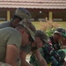 A group of military personnel in mixed camouflage uniforms are collaboratively laying interlocking paving stones or bricks on a sand base. The work appears to be a joint engineering or civic action project, with soldiers using rubber mallets to set the pavers in place. The setting appears to be near a building, suggesting a construction or infrastructure improvement project.