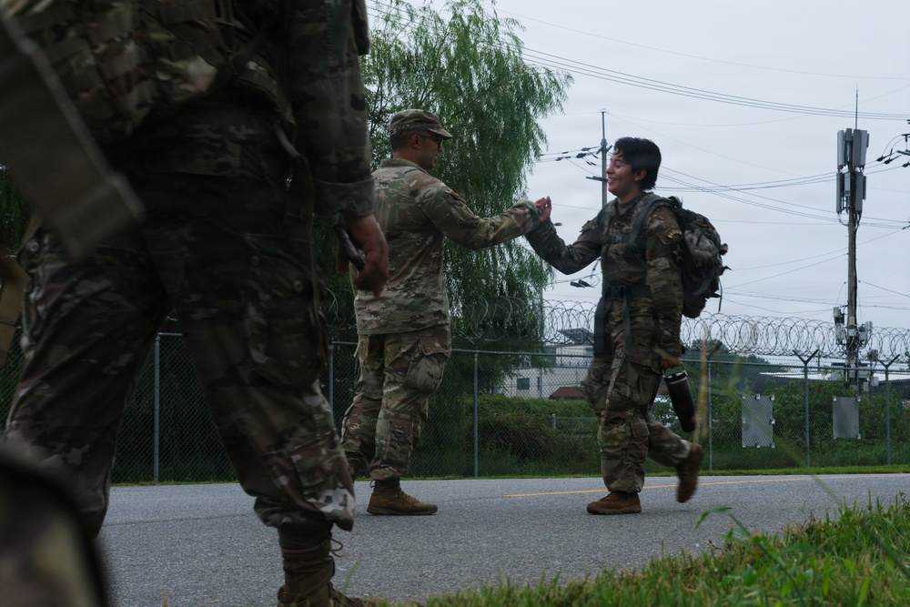 Two soldiers in camouflage uniforms are greeting each other with a handshake or fist bump on a paved road near a military installation. A third soldier is visible in the foreground, partially out of frame. The setting appears to be a secured perimeter area with chain-link fencing topped with razor wire.