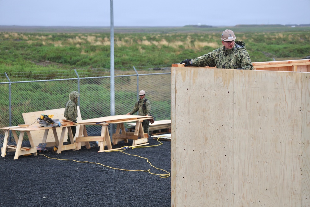 Military personnel in camouflage uniforms and hard hats are engaged in construction work, assembling a wooden structure using plywood panels and lumber. The scene appears to be an outdoor construction site surrounded by a chain-link fence with flat, grassy terrain visible in the background. Multiple workers are visible, with one elevated on the structure and others working at sawhorses with power tools.