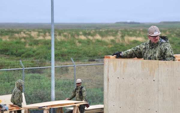 Military personnel in camouflage uniforms and hard hats are engaged in construction work, assembling a wooden structure using plywood panels and lumber. The scene appears to be an outdoor construction site surrounded by a chain-link fence with flat, grassy terrain visible in the background. Multiple workers are visible, with one elevated on the structure and others working at sawhorses with power tools.