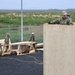 Military personnel in camouflage uniforms and hard hats are engaged in construction work, assembling a wooden structure using plywood panels and lumber. The scene appears to be an outdoor construction site surrounded by a chain-link fence with flat, grassy terrain visible in the background. Multiple workers are visible, with one elevated on the structure and others working at sawhorses with power tools.