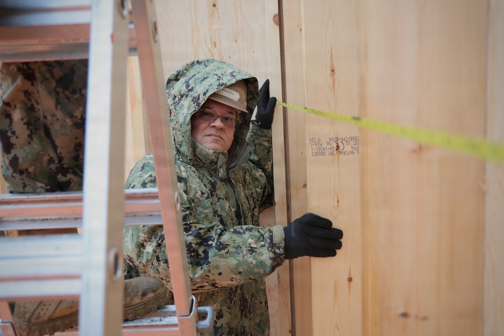 A military service member wearing digital camouflage uniform with hood up and black gloves is working on a wooden structure, appearing to measure or align plywood panels. A yellow measuring tape is visible stretched across the plywood surface. The setting appears to be a construction or building project in cold or wet weather conditions.