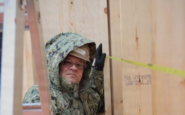 A military service member wearing digital camouflage uniform with hood up and black gloves is working on a wooden structure, appearing to measure or align plywood panels. A yellow measuring tape is visible stretched across the plywood surface. The setting appears to be a construction or building project in cold or wet weather conditions.