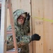 A military service member wearing digital camouflage uniform with hood up and black gloves is working on a wooden structure, appearing to measure or align plywood panels. A yellow measuring tape is visible stretched across the plywood surface. The setting appears to be a construction or building project in cold or wet weather conditions.
