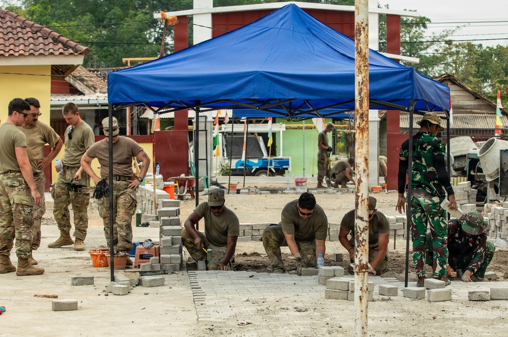 Military personnel from what appear to be two different nations are collaborating on a construction/civic action project, laying interlocking concrete pavers on a prepared sand base. The work is being conducted under a blue pop-up canopy tent at what appears to be a community or school facility. Some soldiers are actively laying bricks while others observe and supervise.