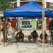 Military personnel from what appear to be two different nations are collaborating on a construction/civic action project, laying interlocking concrete pavers on a prepared sand base. The work is being conducted under a blue pop-up canopy tent at what appears to be a community or school facility. Some soldiers are actively laying bricks while others observe and supervise.