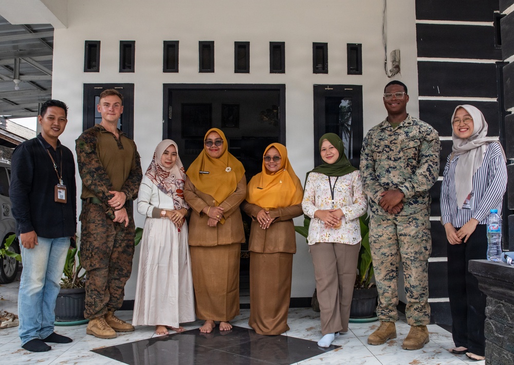 A group of eight people pose for a formal group photo in front of a black-and-white building. The group includes two individuals in U.S. Marine Corps camouflage uniforms and six civilians, several of whom are wearing hijabs, suggesting a community engagement or diplomatic visit in a Southeast Asian country. The setting and attire indicate a cross-cultural military-civilian interaction.