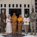 A group of eight people pose for a formal group photo in front of a black-and-white building. The group includes two individuals in U.S. Marine Corps camouflage uniforms and six civilians, several of whom are wearing hijabs, suggesting a community engagement or diplomatic visit in a Southeast Asian country. The setting and attire indicate a cross-cultural military-civilian interaction.