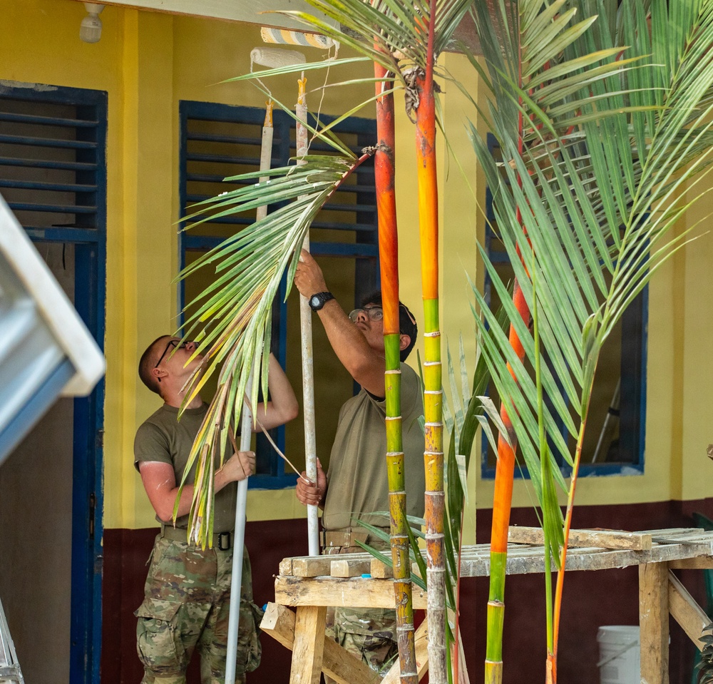 Two individuals wearing military-style olive/camouflage uniforms are working on what appears to be a construction or renovation project at a yellow building with blue-trimmed shutters and doors. They are using long-handled tools, possibly paint rollers or scrapers, while navigating around tall tropical palm plants with distinctive orange and green stalks. The setting appears to be a tropical or subtropical location, likely a community building or school.