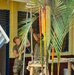 Two individuals wearing military-style olive/camouflage uniforms are working on what appears to be a construction or renovation project at a yellow building with blue-trimmed shutters and doors. They are using long-handled tools, possibly paint rollers or scrapers, while navigating around tall tropical palm plants with distinctive orange and green stalks. The setting appears to be a tropical or subtropical location, likely a community building or school.