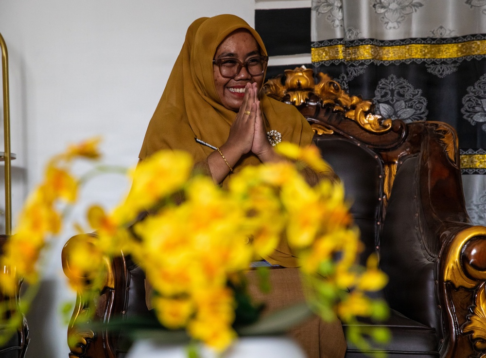 A woman wearing a mustard-yellow hijab and matching outfit is seated in an ornate wooden armchair with gold detailing, performing a traditional greeting gesture with her hands pressed together (salam/namaste). She is smiling warmly and wearing glasses, a brooch, and a gold bracelet. Yellow flowers in the foreground are intentionally blurred using shallow depth of field.