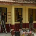 Military personnel in camouflage uniforms are performing renovation or maintenance work on the exterior of a building with yellow and dark red walls. The work appears to involve painting window frames blue and conducting general repairs. Two ladders are set up and multiple workers are engaged at different windows along the building facade.