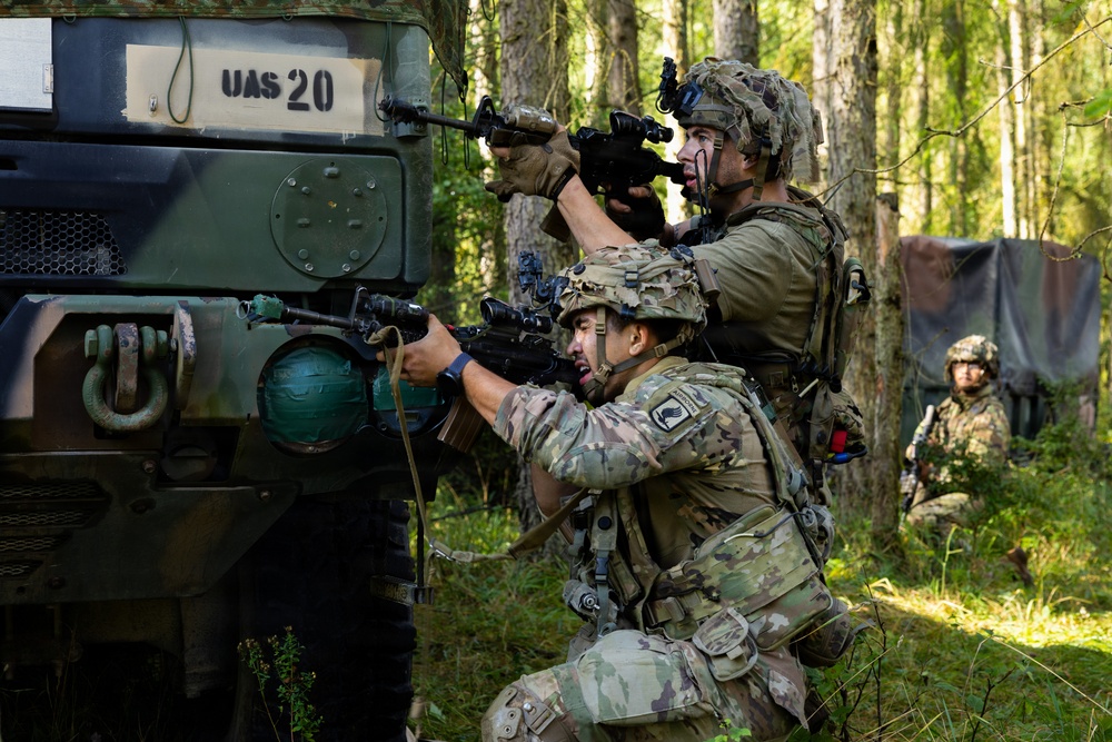 Two foreground soldiers in MultiCam/OCP camouflage uniforms take covered firing positions beside a military vehicle marked 'UAS 20' in a forested environment. Both are armed with rifles equipped with optics and accessories, with one standing and one kneeling. A third soldier in different camouflage pattern is visible in the background near a camouflage tarp.