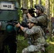 Two foreground soldiers in MultiCam/OCP camouflage uniforms take covered firing positions beside a military vehicle marked 'UAS 20' in a forested environment. Both are armed with rifles equipped with optics and accessories, with one standing and one kneeling. A third soldier in different camouflage pattern is visible in the background near a camouflage tarp.