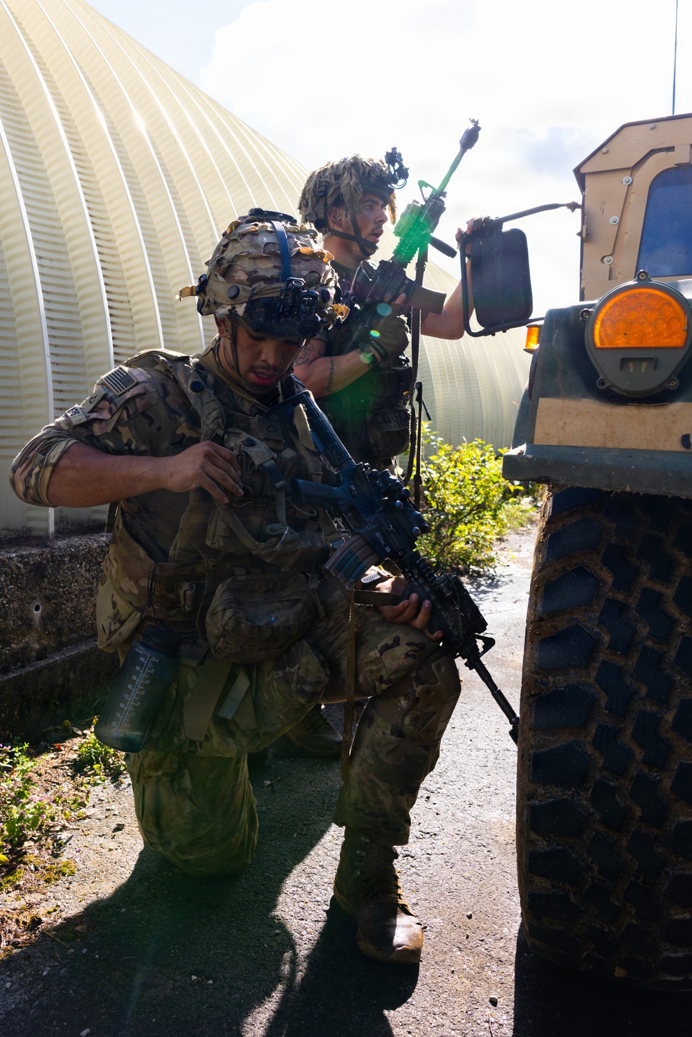 Two soldiers in full combat gear are taking cover beside a tan military HMMWV (Humvee) during what appears to be a training exercise. Both wear MultiCam/OCP camouflage uniforms with tactical helmets and are carrying rifles equipped with training accessories, including a visibly green-colored training device on one weapon. The setting includes a corrugated metal structure in the background and a paved surface.