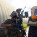 Two soldiers in full combat gear are taking cover beside a tan military HMMWV (Humvee) during what appears to be a training exercise. Both wear MultiCam/OCP camouflage uniforms with tactical helmets and are carrying rifles equipped with training accessories, including a visibly green-colored training device on one weapon. The setting includes a corrugated metal structure in the background and a paved surface.