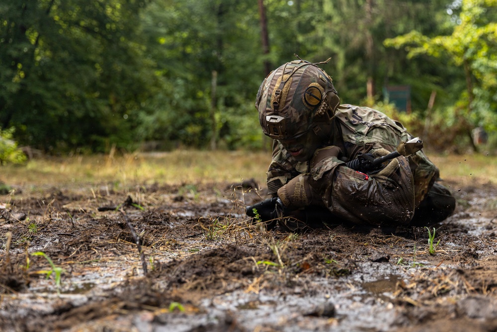A soldier in full camouflage gear and tactical helmet is crouched low in muddy terrain, appearing to examine or interact with something on the ground. The setting is a wooded or forest-edge environment with wet, churned-up soil suggesting recent rain or intensive activity. The image appears to document a military training exercise or field operation.