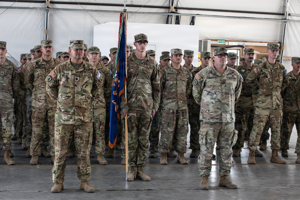 A formation of U.S. Army soldiers standing at attention inside what appears to be a military hangar or large facility. A soldier in the center is holding a unit guidon/flag with blue and gold colors. The formation appears to be part of a formal military ceremony, likely a change of command, deployment, or redeployment event.