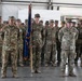 A formation of U.S. Army soldiers standing at attention inside what appears to be a military hangar or large facility. A soldier in the center is holding a unit guidon/flag with blue and gold colors. The formation appears to be part of a formal military ceremony, likely a change of command, deployment, or redeployment event.