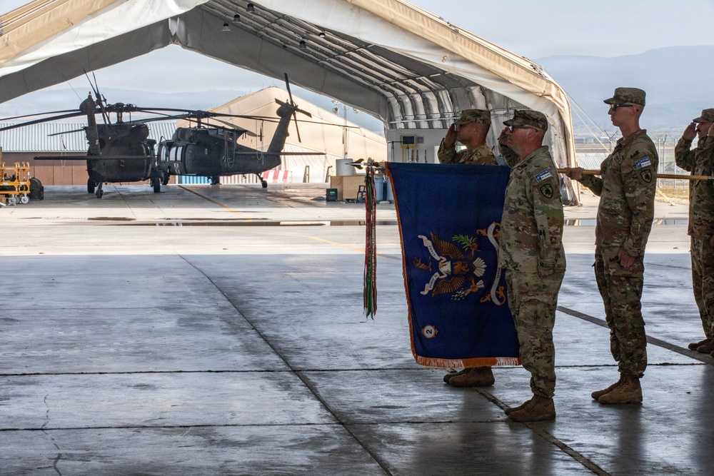 A military ceremony is taking place on an airfield tarmac, with soldiers in OCP (Operational Camouflage Pattern) uniforms rendering salutes while holding a blue unit guidon or organizational flag with gold fringe and streamer decorations. The setting appears to be a forward operating base or military airfield, with UH-60 Black Hawk helicopters visible in the background under a large fabric hangar structure. The scene is consistent with a change of command, activation, or unit casing/uncasing ceremony.
