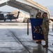 A military ceremony is taking place on an airfield tarmac, with soldiers in OCP (Operational Camouflage Pattern) uniforms rendering salutes while holding a blue unit guidon or organizational flag with gold fringe and streamer decorations. The setting appears to be a forward operating base or military airfield, with UH-60 Black Hawk helicopters visible in the background under a large fabric hangar structure. The scene is consistent with a change of command, activation, or unit casing/uncasing ceremony.