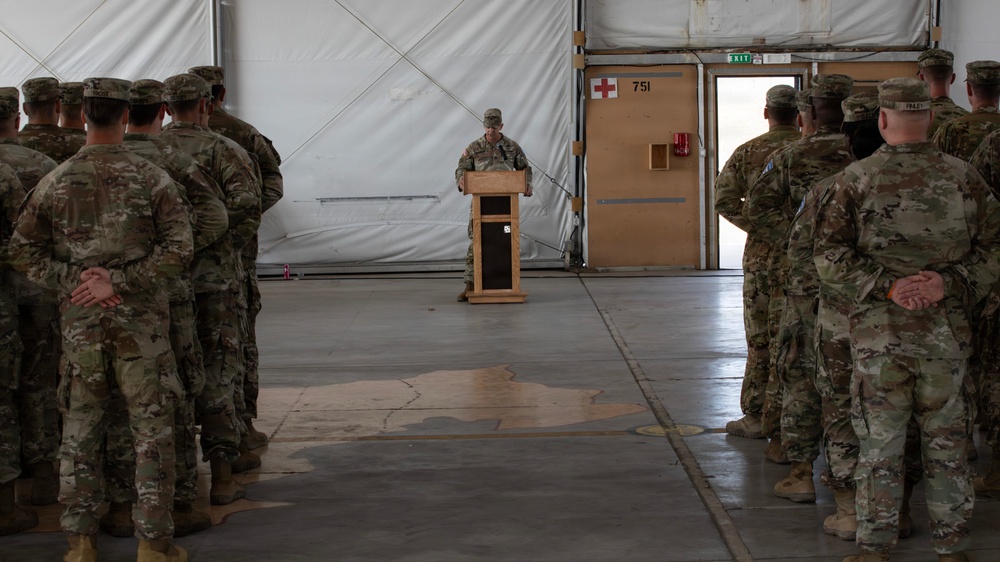 A military ceremony or briefing is taking place inside what appears to be a large hangar or temporary structure. A single soldier stands at a wooden podium addressing two formations of soldiers standing at parade rest on either side. The setting appears to be a forward operating base or deployed location, indicated by the temporary tent-like walls and medical cross signage.