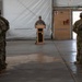 A military ceremony or briefing is taking place inside what appears to be a large hangar or temporary structure. A single soldier stands at a wooden podium addressing two formations of soldiers standing at parade rest on either side. The setting appears to be a forward operating base or deployed location, indicated by the temporary tent-like walls and medical cross signage.