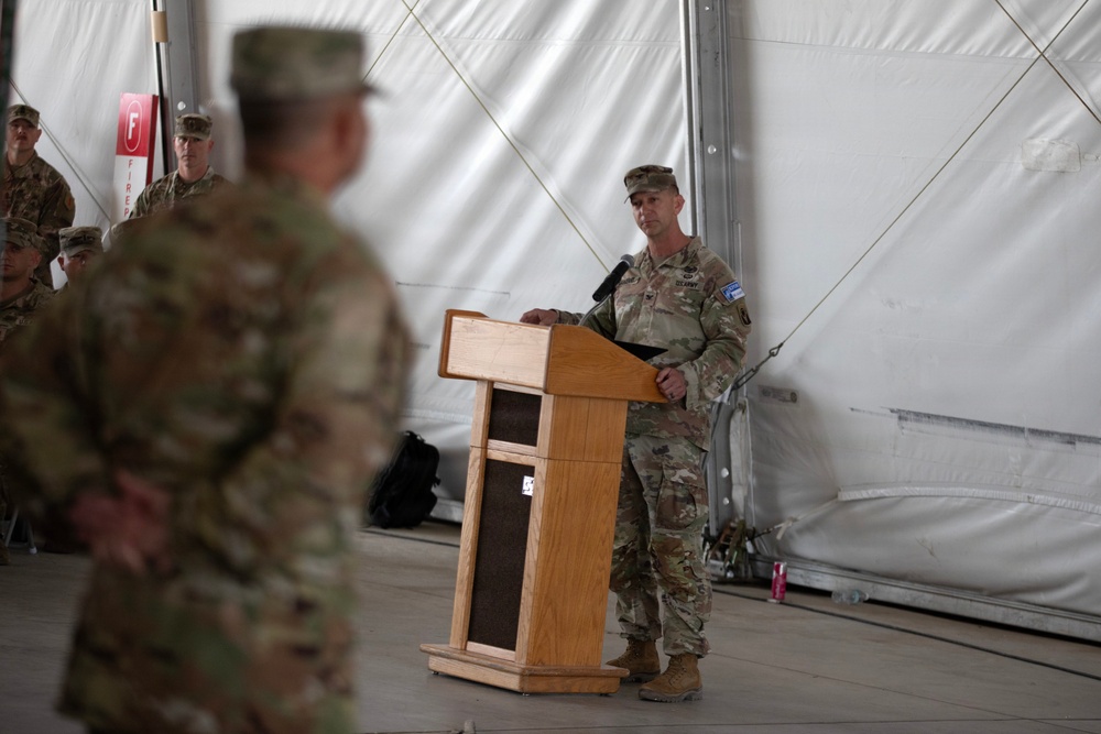 A military ceremony is taking place inside a large tent or temporary structure, with a soldier in U.S. Army combat uniform (OCP/MultiCam) standing at a wooden podium and addressing an audience of fellow soldiers. The setting appears to be a formal military event such as a change of command, award ceremony, or briefing conducted at a forward operating base or military installation. Several uniformed personnel are visible in the background observing the proceedings.
