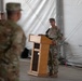 A military ceremony is taking place inside a large tent or temporary structure, with a soldier in U.S. Army combat uniform (OCP/MultiCam) standing at a wooden podium and addressing an audience of fellow soldiers. The setting appears to be a formal military event such as a change of command, award ceremony, or briefing conducted at a forward operating base or military installation. Several uniformed personnel are visible in the background observing the proceedings.
