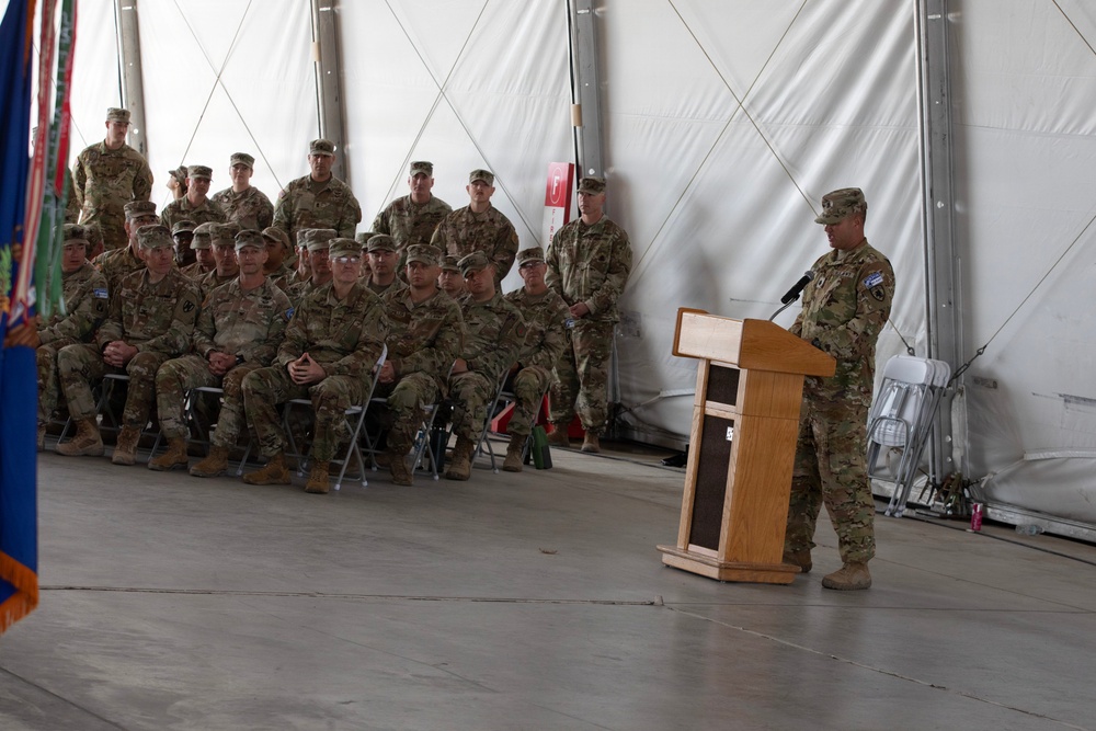 A military ceremony is taking place inside a large tent or temporary structure, with one soldier speaking at a wooden podium while a group of uniformed personnel are seated and standing in formation as an audience. All individuals are wearing U.S. Army combat uniforms (OCP/MultiCam pattern) with patrol caps. The setting appears to be a formal military event such as a change of command, award ceremony, or unit briefing.