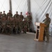 A military ceremony is taking place inside a large tent or temporary structure, with one soldier speaking at a wooden podium while a group of uniformed personnel are seated and standing in formation as an audience. All individuals are wearing U.S. Army combat uniforms (OCP/MultiCam pattern) with patrol caps. The setting appears to be a formal military event such as a change of command, award ceremony, or unit briefing.