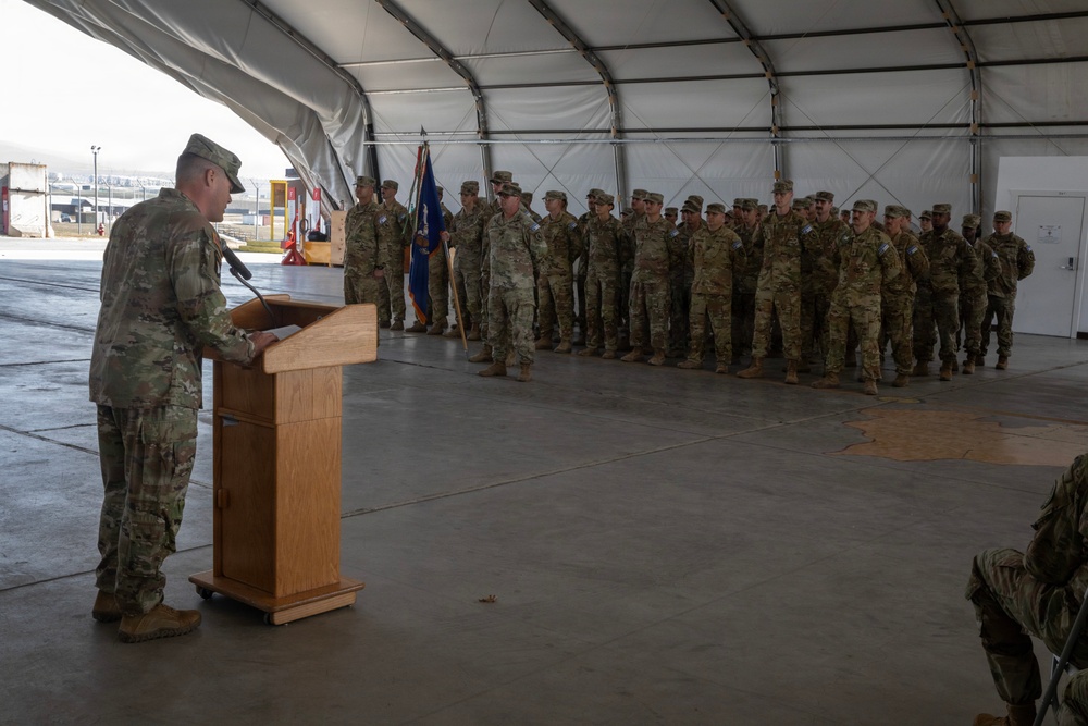 A military officer in OCP (Operational Camouflage Pattern) uniform stands at a wooden podium addressing a formation of soldiers inside a large tent or temporary hangar structure. The assembled troops are standing at attention in rows, with at least one military guidon or unit flag visible among them. The setting appears to be a formal military ceremony, possibly a deployment, redeployment, or change of command event.