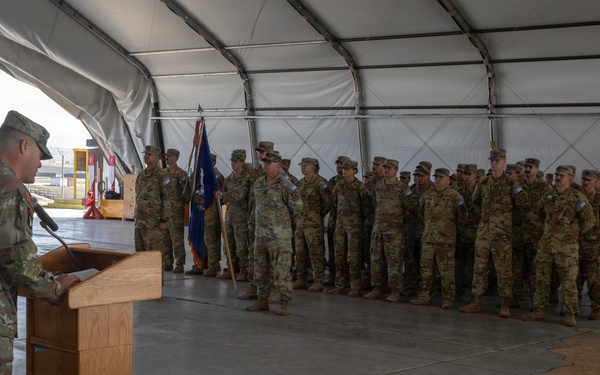 A military officer in OCP (Operational Camouflage Pattern) uniform stands at a wooden podium addressing a formation of soldiers inside a large tent or temporary hangar structure. The assembled troops are standing at attention in rows, with at least one military guidon or unit flag visible among them. The setting appears to be a formal military ceremony, possibly a deployment, redeployment, or change of command event.