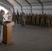 A military officer in OCP (Operational Camouflage Pattern) uniform stands at a wooden podium addressing a formation of soldiers inside a large tent or temporary hangar structure. The assembled troops are standing at attention in rows, with at least one military guidon or unit flag visible among them. The setting appears to be a formal military ceremony, possibly a deployment, redeployment, or change of command event.