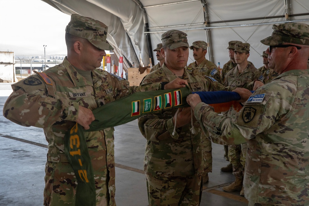 U.S. Army soldiers are participating in a military colors casing ceremony, likely marking the end of a deployment or unit rotation. The soldiers are folding or casing a unit guidon/flag that displays multiple campaign streamers or deployment patches. A KFOR RC-East patch is visible on one soldier's uniform, suggesting a Kosovo Force connection.