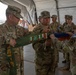 U.S. Army soldiers are participating in a military colors casing ceremony, likely marking the end of a deployment or unit rotation. The soldiers are folding or casing a unit guidon/flag that displays multiple campaign streamers or deployment patches. A KFOR RC-East patch is visible on one soldier's uniform, suggesting a Kosovo Force connection.