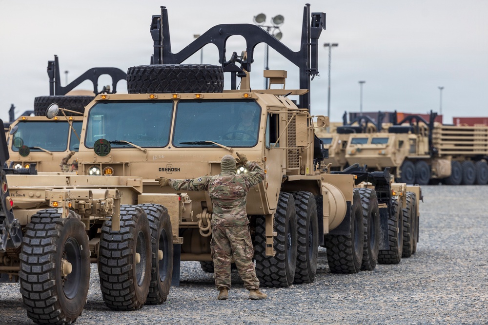 A military soldier in camouflage uniform is directing an Oshkosh heavy tactical vehicle (likely an HEMTT or similar variant) in a staging or marshaling area. Multiple tan/desert-colored military trucks are lined up on a gravel surface, with shipping containers and light poles visible in the background. The scene appears to be a vehicle staging, deployment, or port operations activity.
