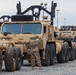 A military soldier in camouflage uniform is directing an Oshkosh heavy tactical vehicle (likely an HEMTT or similar variant) in a staging or marshaling area. Multiple tan/desert-colored military trucks are lined up on a gravel surface, with shipping containers and light poles visible in the background. The scene appears to be a vehicle staging, deployment, or port operations activity.