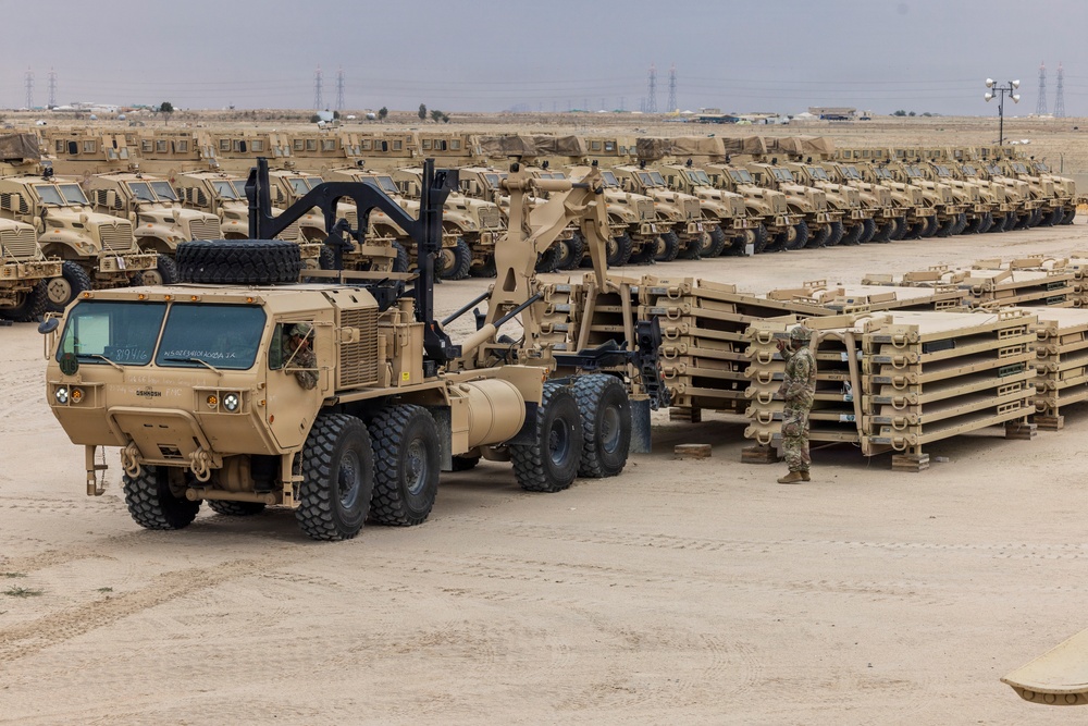 A large military staging area or motor pool in an arid desert environment, featuring rows of tan/sand-colored military vehicles lined up in organized formation. In the foreground, an Oshkosh heavy tactical truck with a crane/loader attachment is positioned near stacked military pallets or flatrack containers. At least one uniformed soldier is visible supervising or conducting logistics operations.